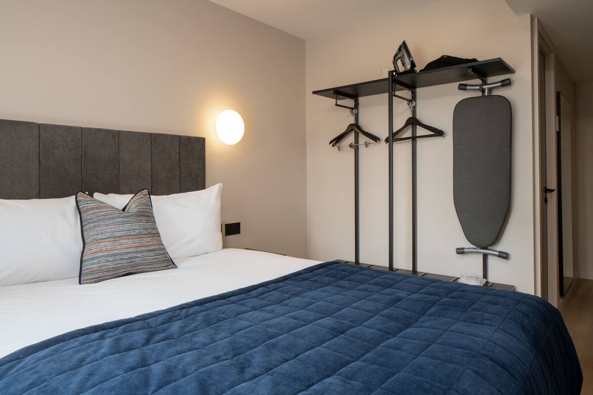Ein ordentlich bezogenes Bett mit einer blauen Steude, einem grauen Kopfteil und einem an der Wand befestigten Bügelbrett mit Kleiderbügeln.