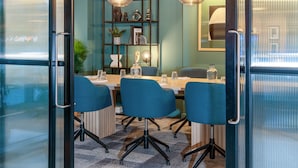 Un salón de conferencias con un panel de vidrio, sillas azules, una mesa de madera y un estante decorativo con diversos objetos.
