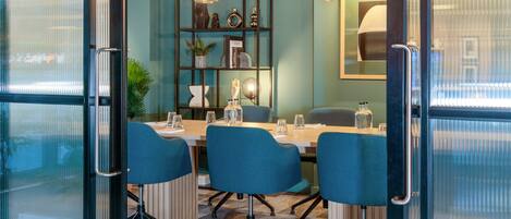 Un salón de conferencias con un panel de vidrio, sillas azules, una mesa de madera y un estante decorativo con diversos objetos.