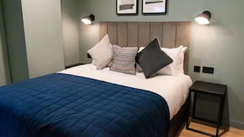 A neatly made bed with a blue quilt, a headboard with vertical panels, two wall-mounted lamps, and a small bedside table.