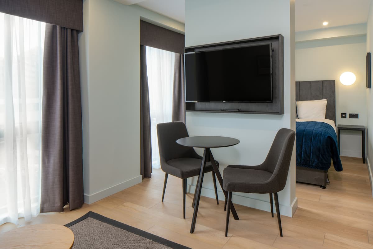 Ein modernes Hotelzimmer mit einem an der Wand befestigten Flachbildfernseher, einem Bett mit blauer Decke, zwei Stühlen um einen kleinen runden Tisch und einem Holzboden.