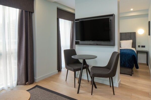 A modern hotel room with a flat-screen TV mounted on the wall, a bed with a blue blanket, two chairs around a small round table, and a wooden floor.