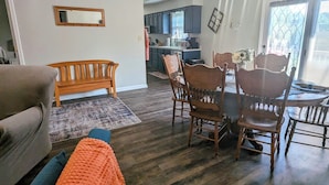 Een woonkamer met een bank, een houten eettafel met stoelen, een keuken op de achtergrond en een groot raam met zonwering.