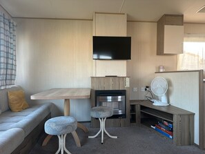 Un espacio habitable compacto con sofá, una mesita, dos taburetes, televisor montado en la pared, un ventilador y una estantería con libros.