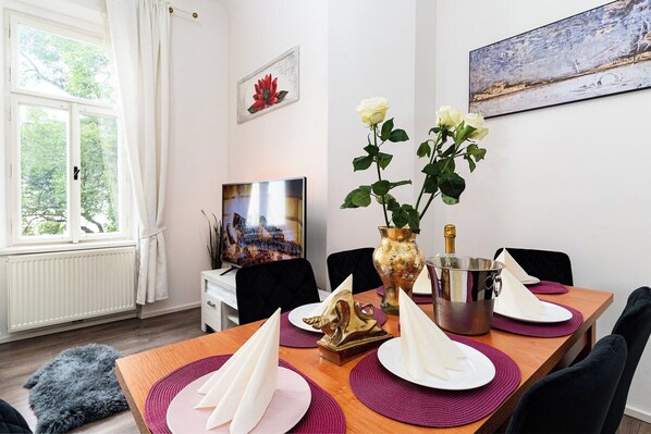 A dining area with a table set for two, a vase with flowers, a champagne bottle, and a painting on the wall.