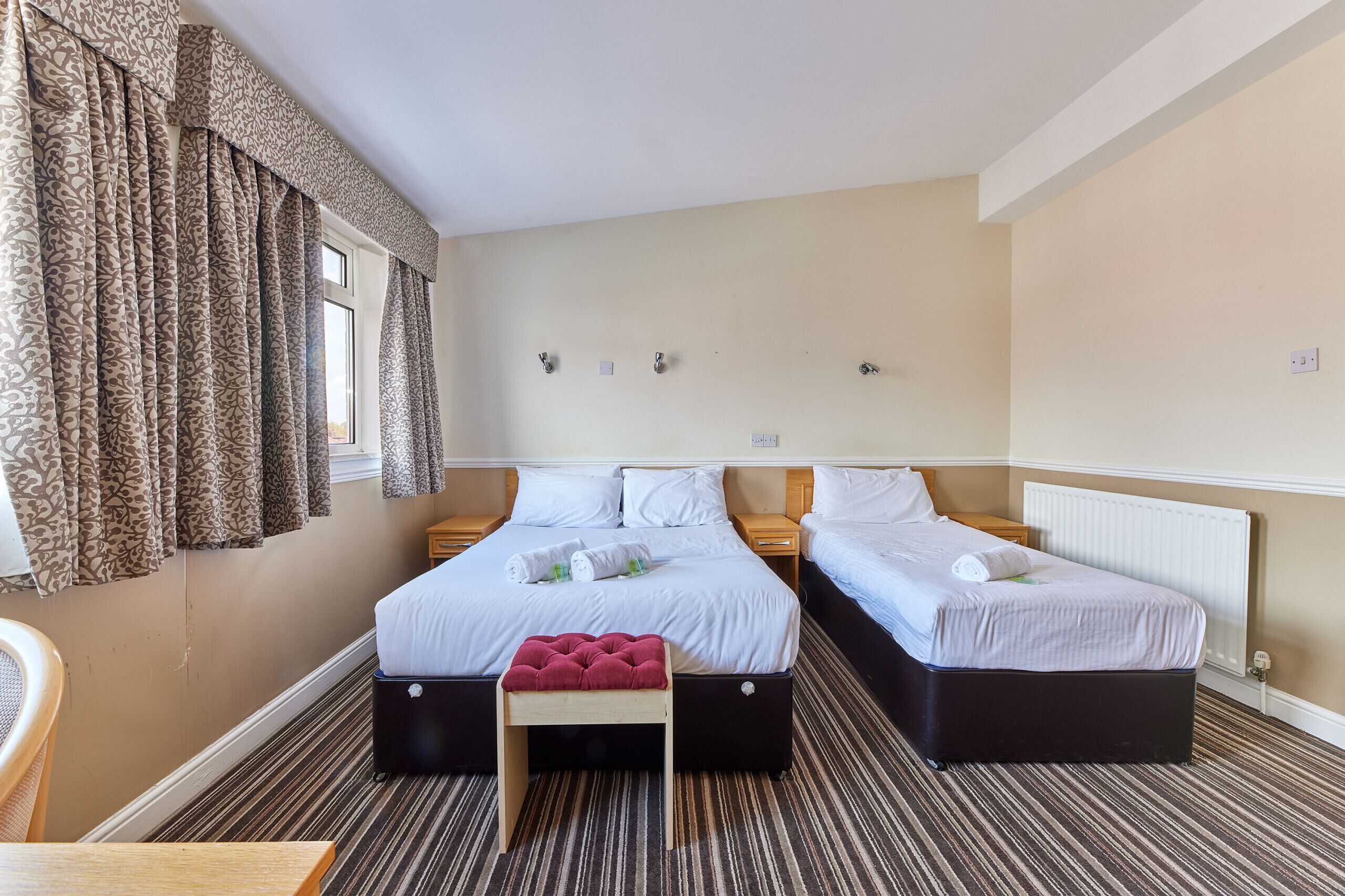 Ein Hotelzimmer mit zwei Betten, einem Fenster mit Vorhängen, einem Streifen Teppich und einem kleinen Holztisch.