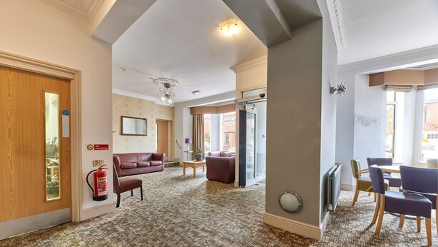 A hotel lobby with a patterned carpet, a fire extinguisher, a sofa, a table, and chairs. - Properties Unique | Dene Rooms Single 38 (Newcastle Upon Tyne)