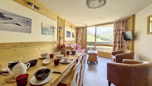 A cozy room with wooden walls, a dining table set with tea cups and saucers, a sofa, and a TV mounted on the wall. - Ski-in/ski-out & foot of the slopes, 6th floor, balcony, tv, ski locker, 40m², Les Menuires (Les Menuires)
