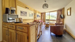 A cozy room with wooden cabinets, a coffee machine, a television, and a view of the outdoors. - Ski-in/ski-out & foot of the slopes, 6th floor, balcony, tv, ski locker, 40m², Les Menuires (Les Menuires)