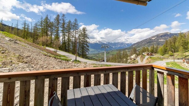Un pontile in legno con tavolo e sedie, affaccia su un paesaggio montuoso con una strada e un piccolo edificio.