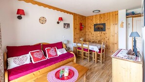 Una stanza accogliente con una parete in legno, un divano rosso con cuscini decorati, un tavolo da pranzo con sedie e un angolo cottura con una lampada.