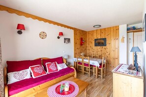 A cozy room with a wooden wall, a red sofa with patterned cushions, a dining table with chairs, and a small kitchen area with a lamp.