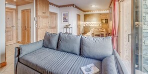 Une pièce chaleureuse avec un canapé gris, des portes en bois, un coin cuisine et une fenêtre avec des rideaux.