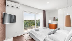 A modern bedroom with a large bed, a flat-screen TV, a desk, and a view of greenery outside.