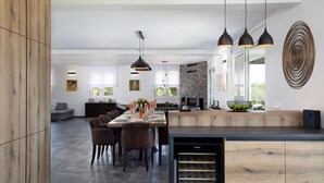 A modern dining area with a long table, multiple chairs, pendant lights, and a kitchen in the background.