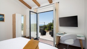 A bedroom with a bed, a desk, a chair, a TV, and a sliding glass door leading to an outdoor area with a view of the sea.