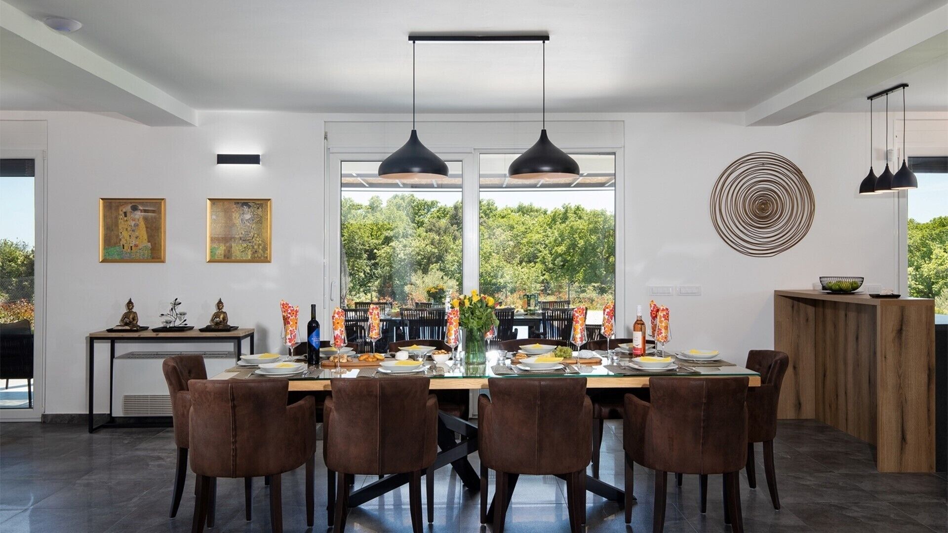 A modern dining area with a large table set for six, surrounded by brown chairs. There are two pendant lights above the table, and a decorative wall piece to the right.