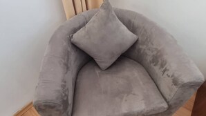 Un fauteuil gris, recouvert de tissu, agrémenté d’un coussin, placé contre un mur orné de stores verticaux.