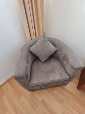 Un fauteuil gris, recouvert de tissu, agrémenté d’un coussin, placé contre un mur orné de stores verticaux.