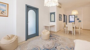 Una habitación de hotel moderna con una cama grande, una zona de estar con un sillón de saco, una mesa de comedor con sillas, y un pasillo con un espejo y obras de arte.