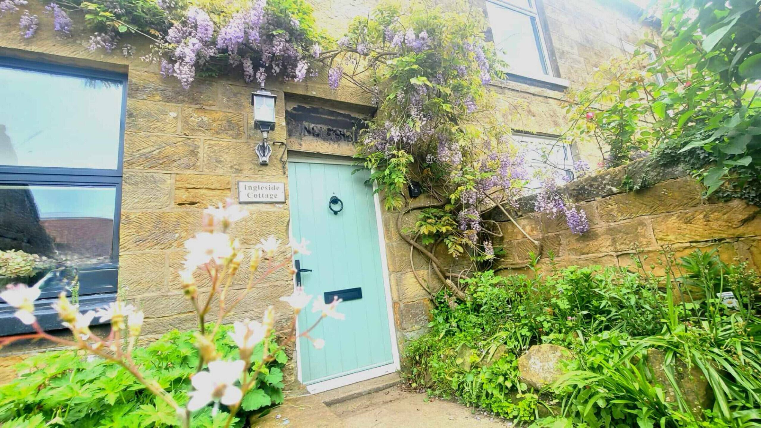 A stone cottage with a light blue door, a sign reading 'Ingleside Cottage,' and a small garden with various plants and flowers.