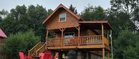 A wooden cabin with a balcony, surrounded by a grassy yard and trees.