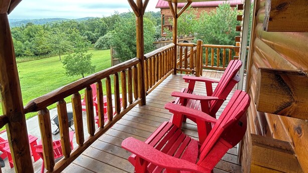 Una terraza de madera con sillas Adirondack rojas, barandilla de madera y vista a un paisaje verde.