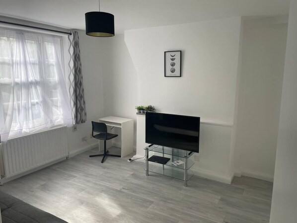 A modern, minimalistic room with a white desk, a black chair, a glass TV stand, and a pendant light.