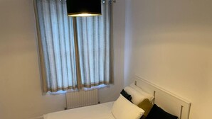 Una camera da letto con un letto, tende bianche, un termosifone e un comodino con una lampada.