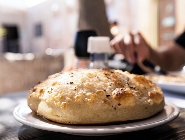 A round, golden-brown baked item on a white plate, possibly a bread or pastry, with a blurred background suggesting a dining setting.