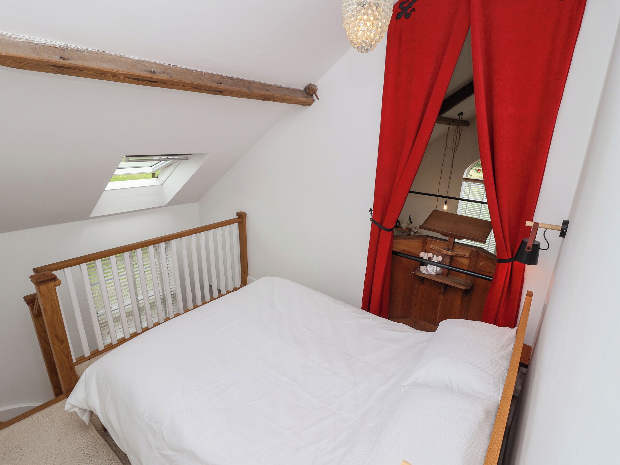 Un dormitorio con techo inclinado, tragaluz, cama con ropa blanca, cabecero de madera, cortina roja y cómoda de madera con espejo.