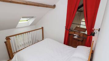 Un dormitorio con techo inclinado, tragaluz, cama con ropa blanca, cabecero de madera, cortina roja y cómoda de madera con espejo.