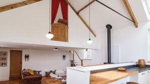A spacious loft with exposed wooden beams, a high ceiling, a kitchen area with a wooden countertop and stools, a cozy seating area with a sofa and cushions, and a red curtain. - The Chapel (Alfreton)
