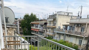 A balcony with a wooden bench and a metal table, overlooking a residential area with buildings and greenery. - Modern, Centrally Located Apartment (Thessaloniki)