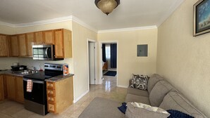A kitchen with wooden cabinets, a microwave, stove, and a sink. There is a living room with a couch and a door leading to another room. - Rolleville Darling House 1 Bedroom Unit (Rolleville)