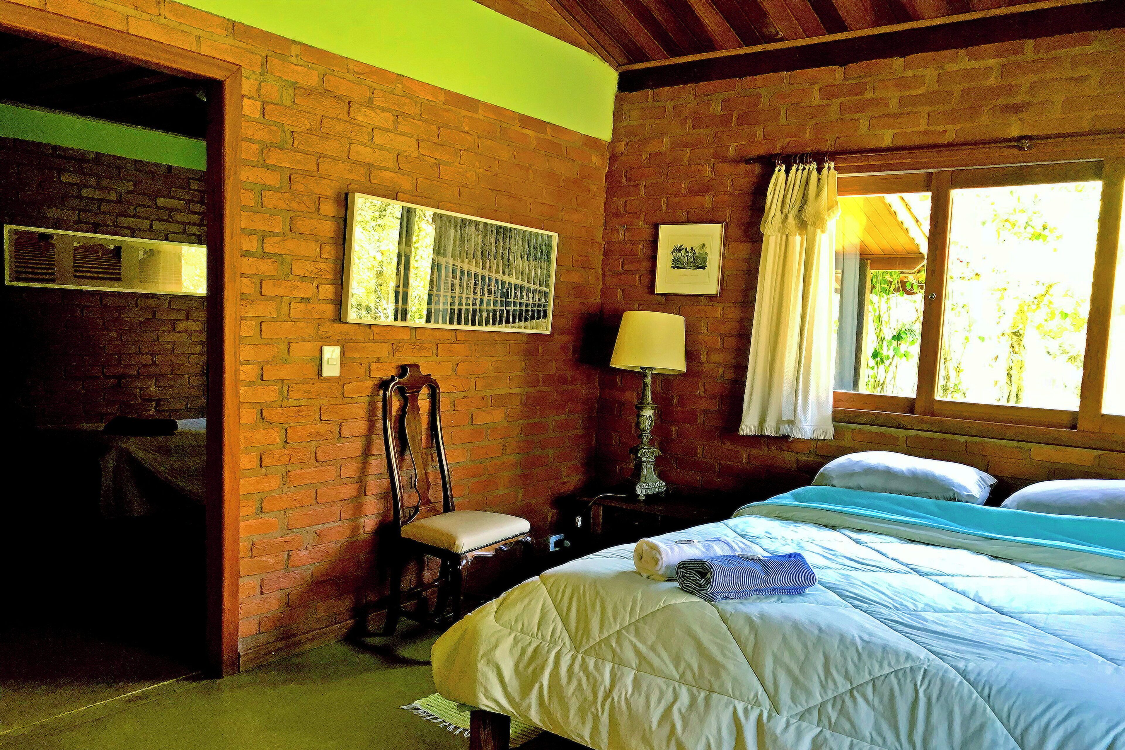 A room with a brick wall, a bed with a quilt, a chair, a nightstand with a lamp, and a window with curtains.