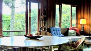 A room with a round table, a blue cushioned chair, a wooden bench, and a view of lush greenery through large windows. - Pousada Sitio Gan Eden (Itatiaia)