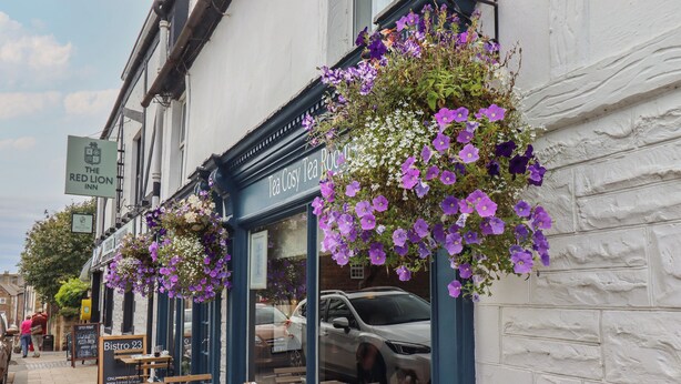 Un espace extérieur aménagé avec des tables et des chaises, une enseigne indiquant « Tea Cozy Tea Room », et des paniers de fleurs suspendus.