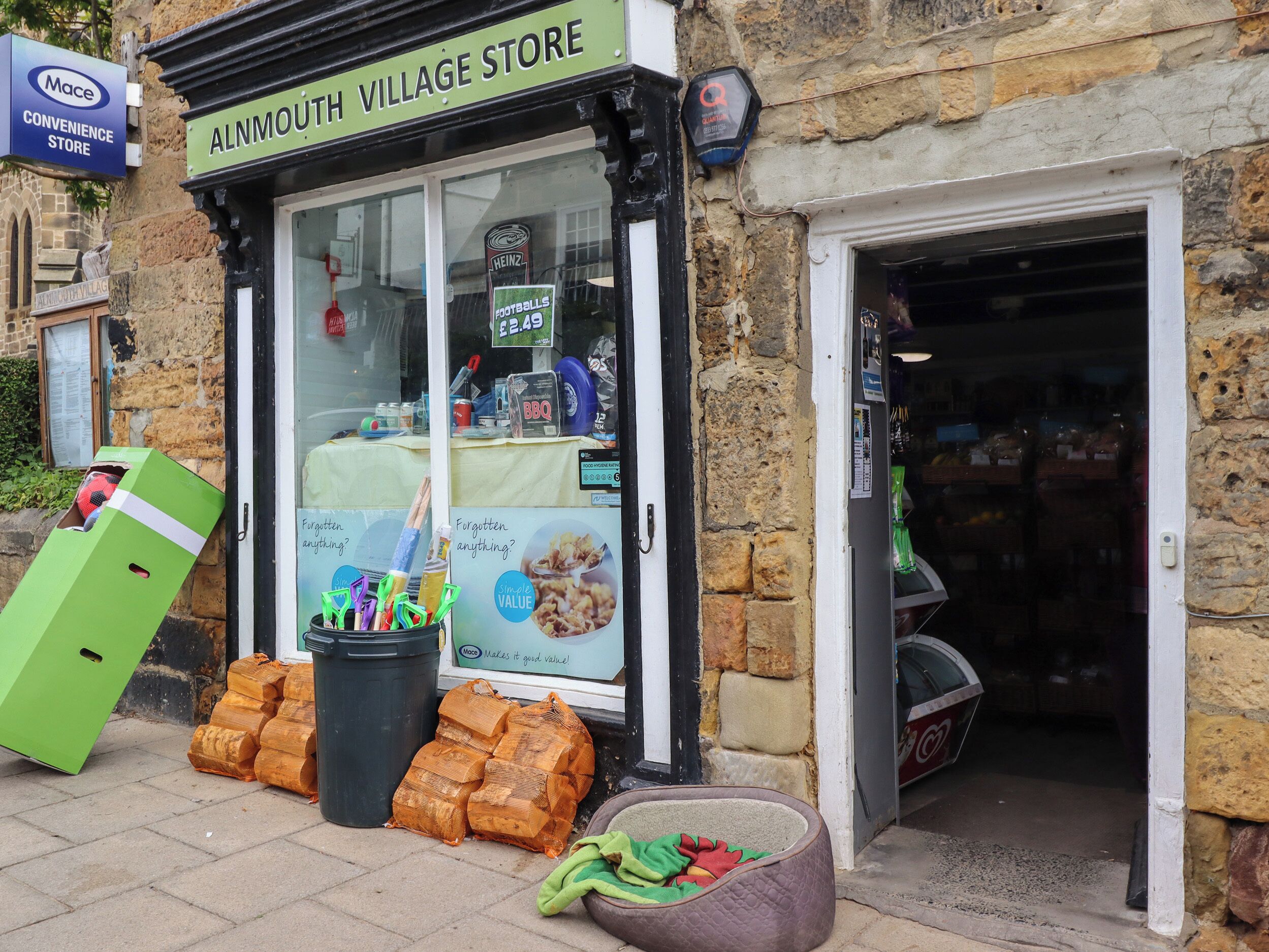 Fachada com uma placa indicando "ALNMOUTH VILLAGE STORE" e diversos itens expostos, incluindo uma caixa verde, uma lixeira preta e uma cama para animais de estimação.