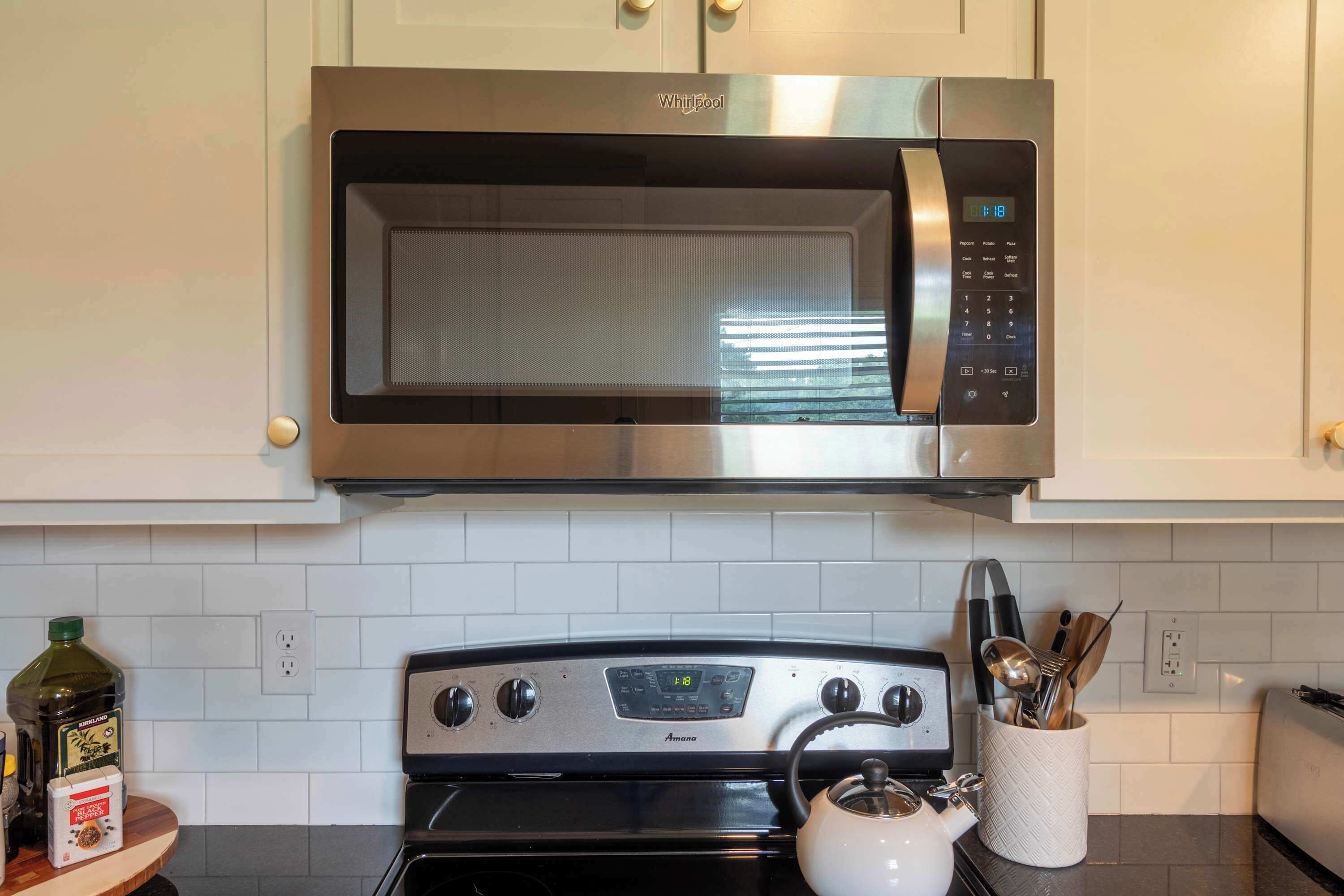 A kitchen with a stainless steel microwave above a stove, a white kettle, and a container with utensils.
