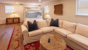 A cozy living room with a beige sectional sofa, a round woven ottoman, a red patterned rug, a wooden side table with a lamp, and a bunk bed in the background. - 4 BR Sleeps 10 Near UGA & Downtown (Athens)
