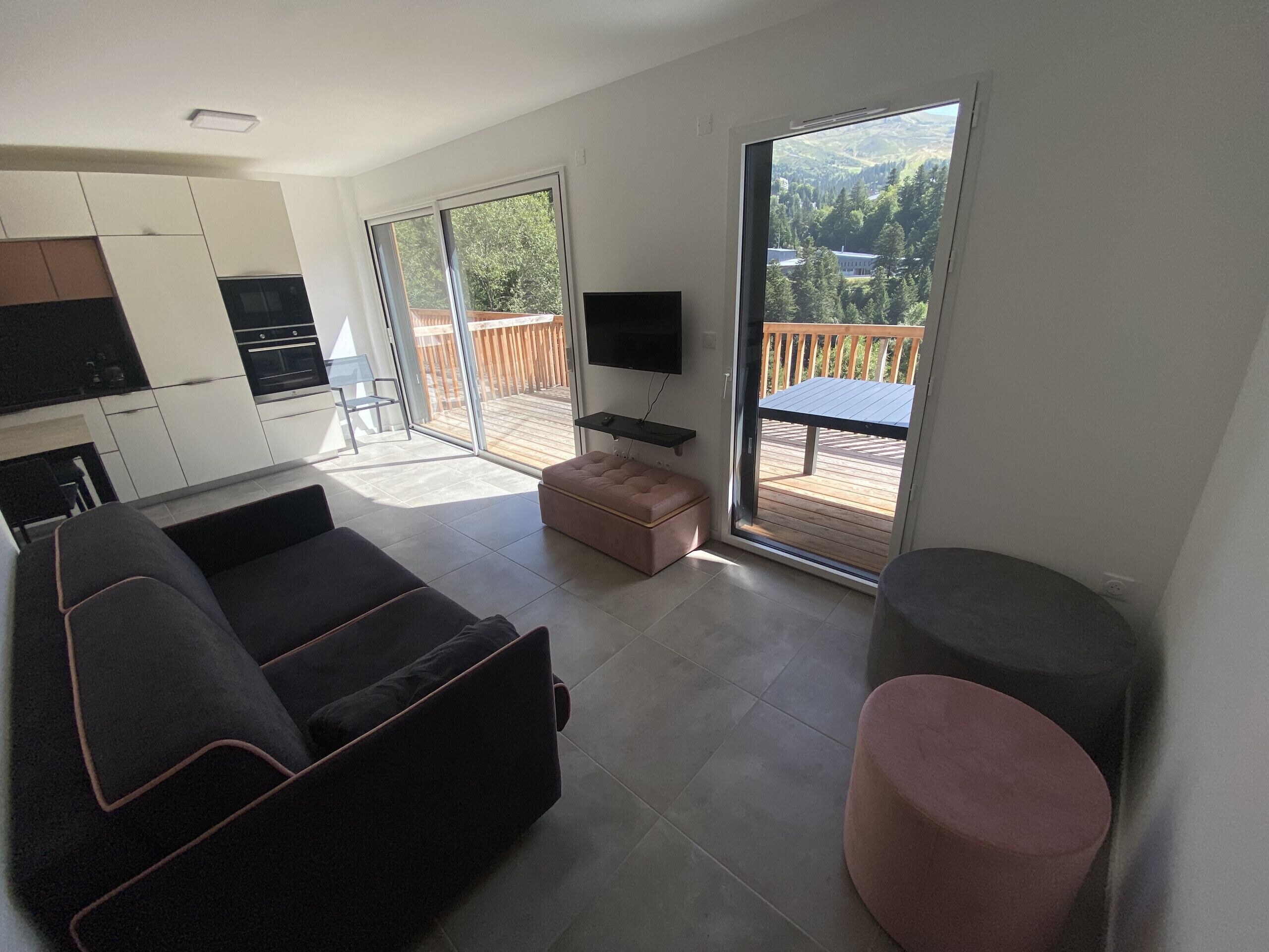 Een moderne woonkamer met een bank, een poef en een televisie op een standaard. Er is een schuifdeur naar een balkon met een houten terras.
