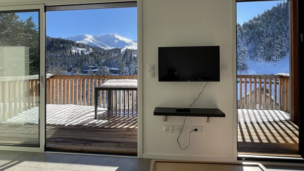 Une pièce avec un téléviseur fixé au mur, une terrasse en bois offrant une vue sur les montagnes enneigées et une porte-fenêtre coulissante donnant sur la terrasse.
