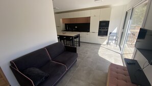 Un salon moderne avec un canapé foncé et un pouf rose, un coin repas avec une table et des chaises, et une cuisine avec des armoires blanches et un plan de travail noir.