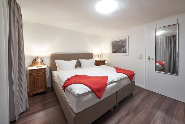 A neatly made bed with a red throw blanket, a wooden nightstand with a lamp, a framed picture on the wall, and a mirror on the door.
