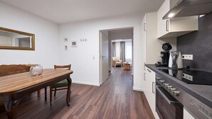 Ett modernt kök med en inbyggd ugn och en kaffebryggare, en matplats med ett träbord och stolar, och en hall som leder till ett vardagsrum med en soffa och gardiner.