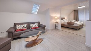 Ett modernt, väl upplyst rum med en soffa, ett soffbord med glasskiva och en säng med ett nattduksbord.