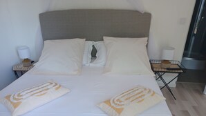 Un lit bien fait, avec du linge de lit blanc et des coussins décoratifs, une table de chevet avec une lampe et un panier, et un espace cuisine moderne visible à travers une porte ouverte.