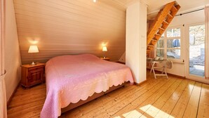 A bedroom with a wooden floor, a bed with a pink quilt, two bedside lamps, a wooden ladder, and a window with a view of the outside.