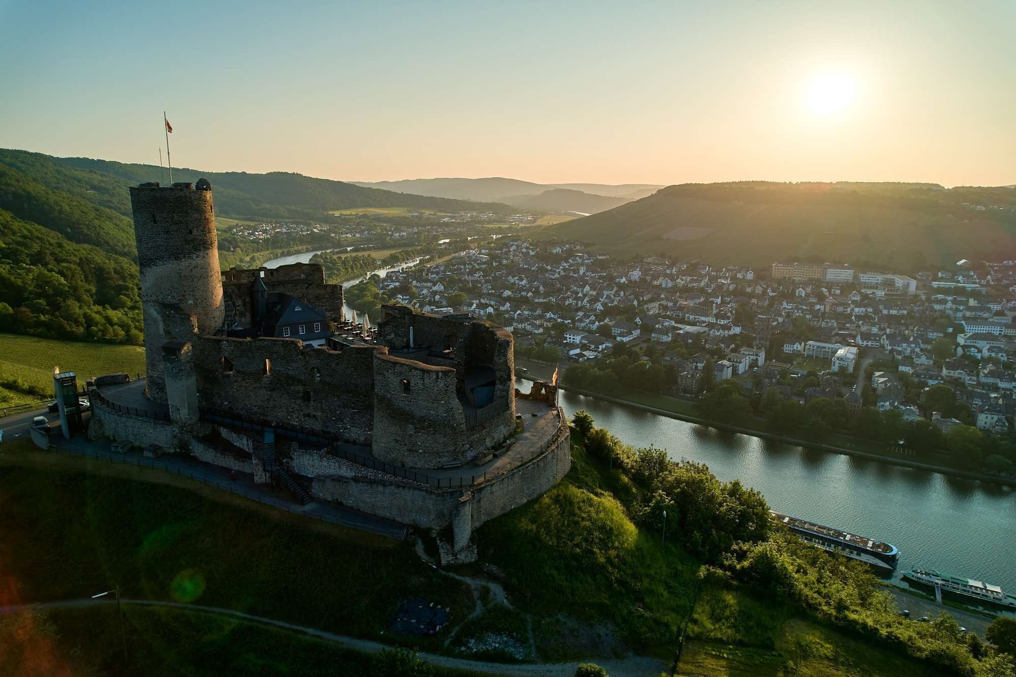 A historic castle with a river in the background, surrounded by greenery and a town.